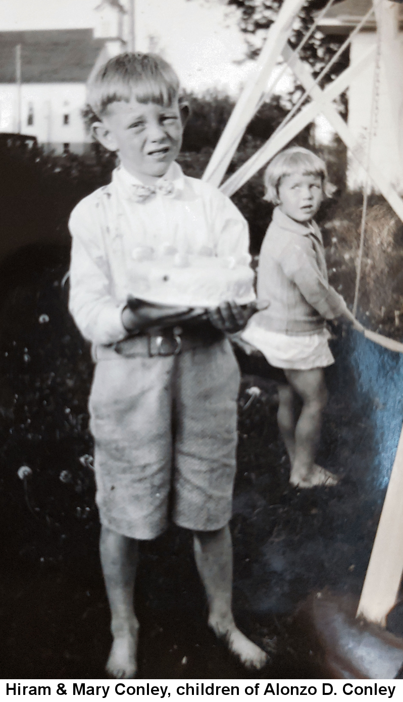 Black and white photo of tow-headed Hiram Conley, about six years old, barefoot and in sorts and white shirt, holding a white-frosted and decorated cake carefully in both hands. His sister, about three years old, with short blonde hair, wearing a long-sleeved sweater and short skirt, also barefoot, stands behind him and to his left. Both are outdoors in a yard dotted with dandelions, standing near a structure with a white frame church in the background.